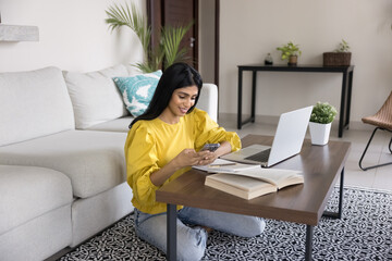 Woman sits on floor at table with books and laptop, using smartphone distracted from studying,...