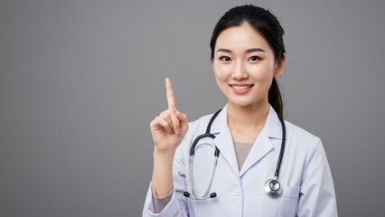 Asian female doctor in white lab coat points upward indoors, promoting health awareness and education
