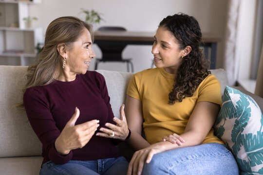 Older 60s Hispanic woman lead pleasant, trustworthy conversation with adult 20s daughter or granddaughter seated on sofa at home. Intergenerational communication, wisdom-sharing, emotional connection - Powered by Adobe