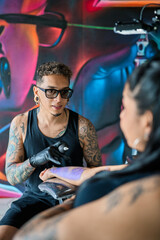Tattoo artist drawing a tattoo on the arm of a female customer in a studio with colorful graffiti on the wall