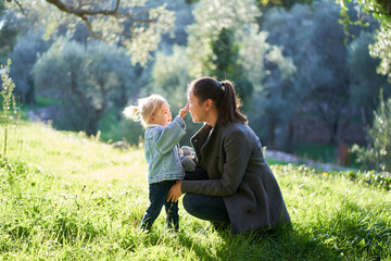 Little girl touches her mom nose while squatting on a green lawn