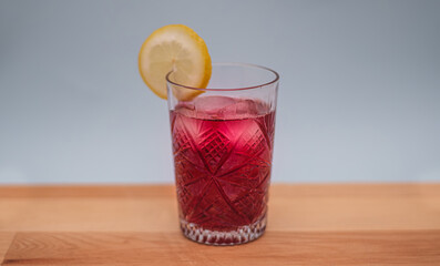 Refreshing iced red beverage with lemon slice on wooden table. An intricately patterned glass. Summer beverages, recipes and lifestyle concepts. Selective focus.