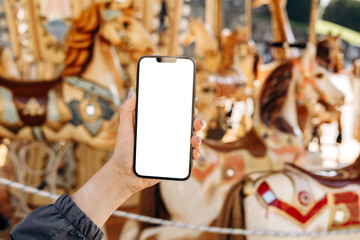 Mockup woman's hand holding using smart phone with white blank screen POV on the background of a carousel with horses, mobile app design and advertising, online marketing © KEA