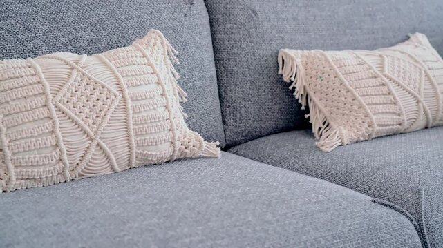 A close-up of a beige macrame pillow with intricate woven patterns and fringed edges resting on a modern gray fabric sofa, adding a cozy and stylish bohemian touch to the decor.