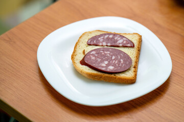 simple sandwich made of bread and a half slice of smoked sausage is placed on a plate on a table. This minimalistic meal reflects a modest dining experience