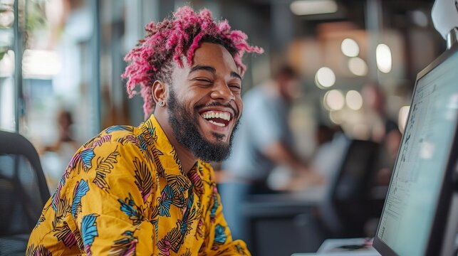 Joyful young man with colorful hair and shirt smiling at computer in modern creative workspace