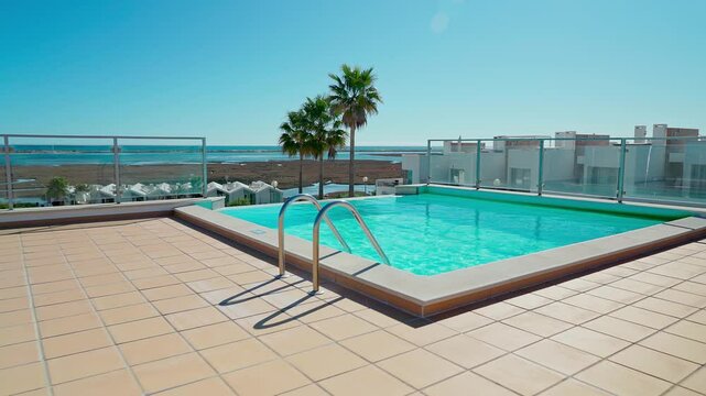 A rooftop swimming pool with clear blue water, surrounded by green and white sun loungers with neatly rolled towels, offering a scenic ocean and palm tree view.