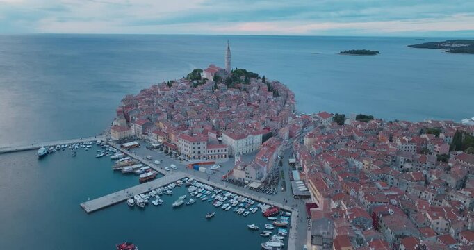 Approaching the historic peninsula of Rovinj in the evening, the drone offers a romantic view of the orange roofs, church towers and the Adriatic Sea. Flying by the port.