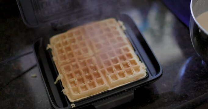 A golden waffle cooking inside a black waffle maker, slightly lifted to reveal the crisp texture. The warm lighting highlights the grid pattern and fresh-baked surface.