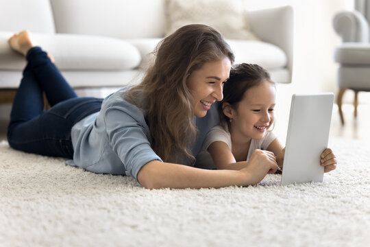Digital family time. Happy bonding young mom little child daughter relax on floor lie on cozy carpet hug look on tablet screen engaged in interactive online learning use web app to play education game