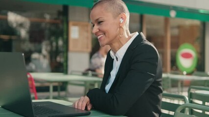 A businesswoman in a suit sits at an outdoor cafe table with her laptop, actively engaged in a video conference using her wireless earbuds.