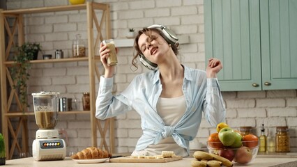 Young woman at the table in the kitchen preparing healthy breakfast, listens music in headphones on smartphone, dancing and drinking smoothie.