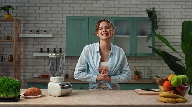 Young woman blogger at the table in the kitchen recording the process of healthy breakfast preparation, talking at camera, showing fruits for fresh smoothie. Slow motion
