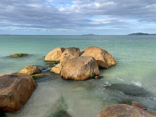 Golden big boulders sunbathed by the late afternoon sun. Brown and orange stones, green turquoise sea, sky full of gray and white clouds, mountains in the horizon. Jurere Internacional beach.
