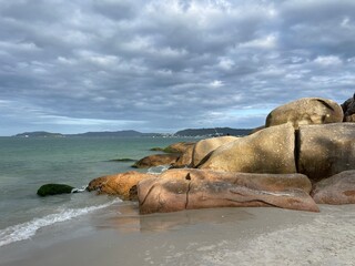 Golden big boulders sunbathed by the late afternoon sun. Brown and orange stones, green turquoise sea, sky full of gray and white clouds, mountains in the horizon. Jurere Internacional beach.