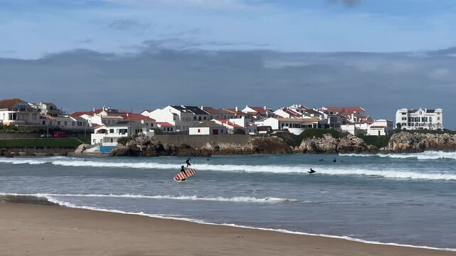 Surfers on the waves at Baleal Norte beach, Portugal. Colorful houses with red roofs line the coast under a blue sky. Peaceful seaside vibe.