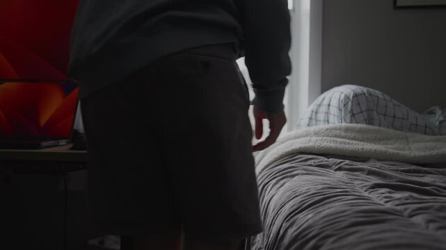 Medium Close-Up Shot of a 20-Year-Old Asian Male Wearing Glasses and a Backward Hat, Opening Window Curtains, Wiping Dust off His Table, and Kneeling to Retrieve Something from Under His Bed
