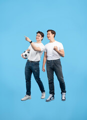 Fototapeta premium Two friends anticipating a soccer match, one holding a ball and pointing, both in casual attire against a blue background. Excitement visible.