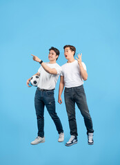 Excited soccer fans pointing. Two young men cheer for their team, one holding a ball against a blue background.
