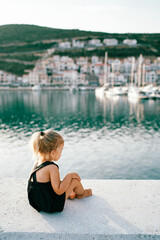Little girl is sitting on the pier and looking at the sea. Side view