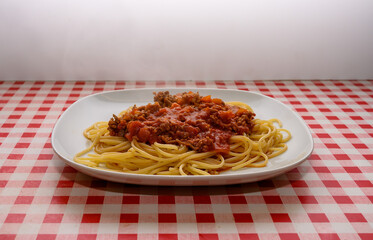 A generous serving of spaghetti topped with rich meat sauce is presented on a white plate. The distinctive red and white checkered tablecloth adds a cozy and inviting feel