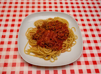 A generous serving of spaghetti topped with rich meat sauce is presented on a white plate. The distinctive red and white checkered tablecloth adds a cozy and inviting feel