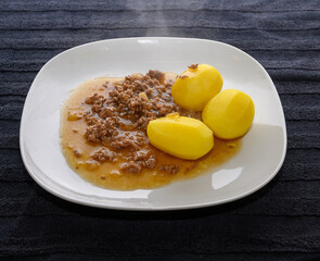 A hearty serving of beef stew sits alongside three perfectly boiled potatoes on a white plate. The dish is steaming and ready to be enjoyed