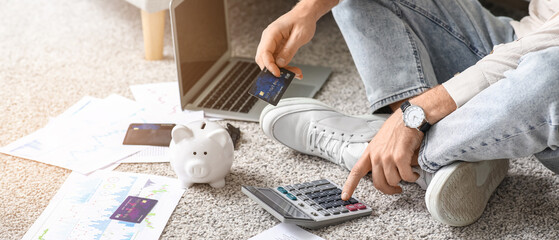 Young man in debt with credit card using calculator on carpet at home, closeup