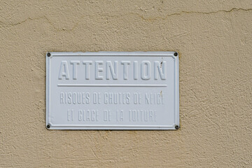 A white metal warning sign with the French words "Attention. Risk of snow and ice falling from the roof" on a yellow wall, Chamonix, France
