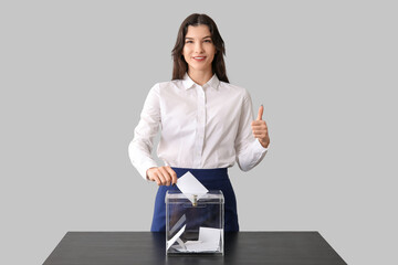 Voting young woman showing thumb-up near ballot box on light background