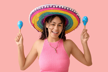Beautiful young African-American woman in sombrero hat with maracas on pink background