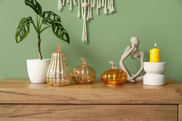 Decorative pumpkins with plant, statuette and burning candle on commode near green wall