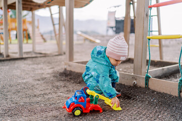 Little girl squats and plays with a toy excavator, shoveling sand