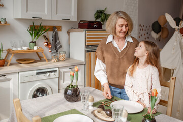Elderly woman grandmother and little granddaughter joyfully set the table at home in the kitchen, learn etiquette