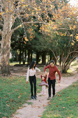 Obraz premium Mom and dad are walking along the path in the park, swinging their little daughter in their arms