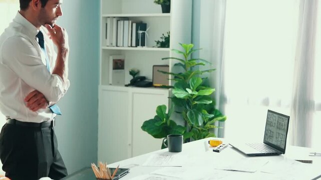 Professional civil engineer standing and drinking coffee and thinking about house plan at meeting table with equipment scatter around. Stressful project manager looking at blueprint. Alimentation.