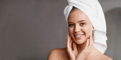 A woman with a towel on her head smiles while pampering her face. She gently massages her skin, embracing self-care and anti-aging practices in a soothing environment, copy space
