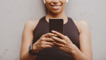 Smart technology for sport. Smiling african american girl with headphones and fitness tracker looks at smartphone on gray wall background, free space