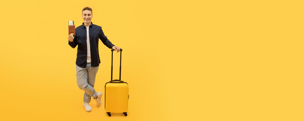 Happy handsome stylish young guy tourist posing with yellow suitcase, showing his cheap flight tickets and passport, isolated on yellow studio background. Travelling, tourism, easy booking