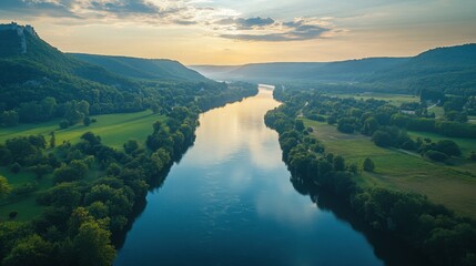 Fototapeta premium River flows between green hills. Water reflects sky light. Landscape shot from above. Nature scene shows peaceful valley. Serene view with trees.