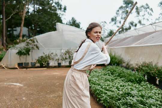 Young woman dressed in an apron, expressing determination while holding a stick in a garden, showcasing resilience in a natural setting with green plants