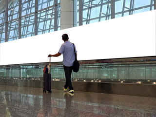 rear view of traveler with suitcase and bags walking in airport hallway while looking at blank white billboard