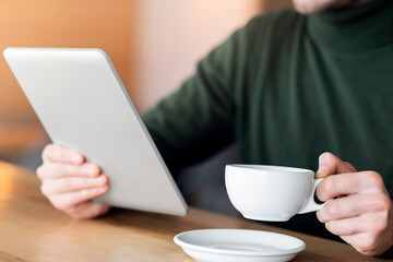 Close up of male hands holding cup of tea and digital tablet, working at cafe