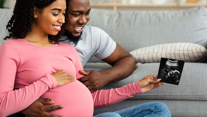 Closeup of smiling african pregnant couple holding sonogram of future baby, living room interior, copy space