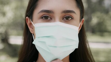 A young Middle Eastern woman stands outdoors in a park, adjusting her face mask with a focused expression. She embodies a modern lifestyle while taking precautionary measures.