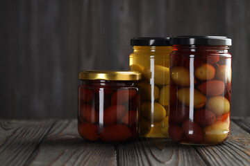 Pickled olives in glass jars on wooden table against gray background, space for text