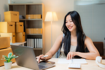 Young asian woman entrepreneur using laptop and taking notes while working in her home office or warehouse, surrounded by cardboard boxes, managing online sales and e-commerce business