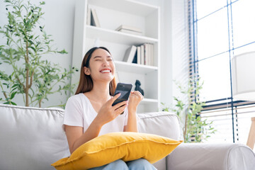 Happy young asian woman relax on comfortable couch at home texting messaging on smartphone, smiling...