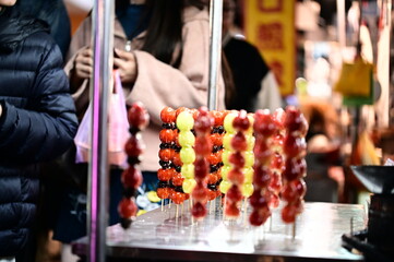 A tanghulu stall at a lively winter night market glows brightly in the cold night. The shiny sugar-coated fruits, such as strawberries, reflect the light beautifully, attracting passersby.
