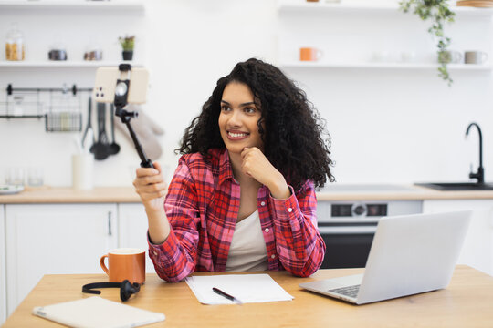 Young African woman recording video on smartphone, sitting in kitchen with laptop and notes. Casual setting for personal or professional content creation